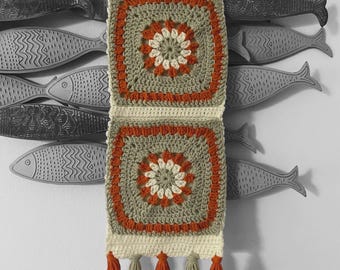 May include: A handmade crochet wall hanging with a square design in shades of orange, cream, and green. The piece features a floral center and is adorned with orange and cream tassels. It hangs from a wooden dowel.