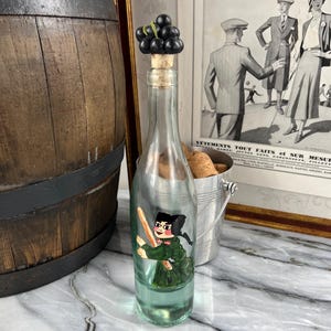 Puede incluir: Una botella de vidrio transparente con un tapón de corcho coronado con un racimo de uvas negras. La botella tiene una figura pintada con un atuendo verde. Un anuncio vintage está en el fondo.