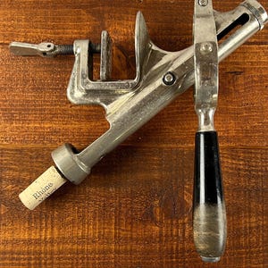 May include: A vintage wine bottle corker made of silver metal with a black handle. The corker is clamped onto a wine bottle cork, which has the word "Rhône" printed on it. The corker is on a wooden surface.
