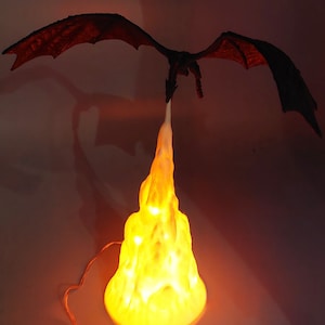 Pode incluir: Uma figura de dragão preto com asas abertas, voando sobre uma base de lâmpada branca em forma de chama. A base da lâmpada está iluminada com um brilho amarelo quente.