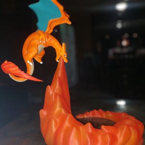 Puede incluir: Figura naranja de Charizard con alas azules y una base en forma de llama. El dragón está en posición de vuelo, con la cola y las alas extendidas. La base es de un naranja vibrante, lo que añade dinamismo a la pieza.