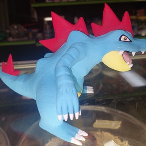 May include: A blue and red Pokemon figurine with a spiky red crest and white claws. The figurine is posed in a standing position with its mouth open.