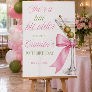 May include: A rectangular white welcome sign on a wooden easel featuring a martini glass illustration with two olives and a pink ribbon. The text reads: She's a tini bit older, WELCOME TO Camila's 30TH BIRTHDAY, and JULY 25, 2027 in pink and green.