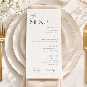 May include: A white rectangular menu card with black text reading The Menu, To Begin, Main, Dessert, Drinks, and the names Emily + Harry with the date 07.11.20XX and New York, NY. The menu is centered on a beige napkin atop a white plate with gold trim.