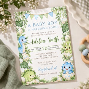 May include: A rectangular baby shower invitation card featuring a dinosaur theme with blue and green illustrations of hatchlings in eggs, tropical leaves, and bunting. The text reads A BABY BOY IS HATCHING SOON, Adeline Smith, October 10, and registry details.