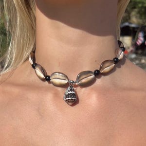 Puede incluir: Un collar gargantilla con grandes conchas marinas marrones y beige, intercaladas con cuentas negras. Un pequeño colgante de concha rayada cuelga del centro. El collar se lleva alrededor del cuello.