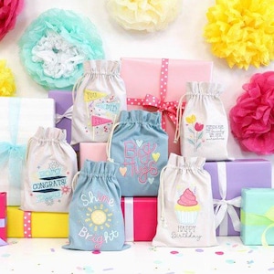 May include: An assortment of gift bags and wrapped presents in various colors, including pink, blue, yellow, and purple. The gift bags feature embroidered designs and text such as "Shine Bright" and "Happy Birthday."