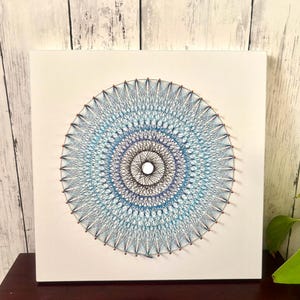 Può includere: Una tela quadrata bianca presenta un design dettagliato di string art. L'opera d'arte presenta un motivo circolare con anelli concentrici di fili blu, turchese e neri che si irradiano da un punto centrale. La string art è montata su una tela bianca.