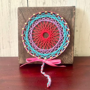 Può includere: Un'opera d'arte con filo colorato con un design circolare con fili rossi, rosa, turchese e viola. L'opera è montata su un blocco di legno scuro e decorata con perline color oro. Un nastro rosa e un cordoncino ritorto sono attaccati in basso.