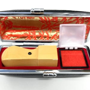 May include: A rectangular black case with a silver metal frame, containing a light-coloured wooden stamp and a square red ink pad. The interior of the case is lined with red velvet and features a gold and red patterned fabric on the lid interior.