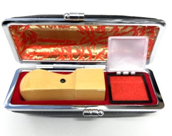 May include: A rectangular black case with a silver metal frame, containing a light-colored wooden stamp and a square red ink pad. The interior of the case is lined with red velvet and features a gold and red patterned fabric on the lid interior.