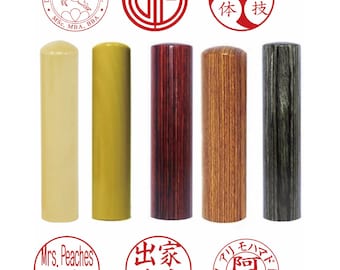 May include: Five cylindrical stamps in varying finishes: cream, mustard yellow, dark reddish-brown wood grain, light brown wood grain, and charcoal grey wood grain. Above and below are six circular red ink impressions featuring text and symbols.