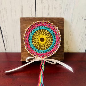 Può includere: Un'opera d'arte colorata con fili che presenta un design circolare con fili gialli, blu e rosa. L'opera è montata su un blocco di legno quadrato e adornata con un nastro bianco e nappe. Un oggetto decorativo.