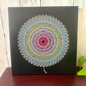 Può includere: Un'opera d'arte colorata con filo su una tela quadrata nera. Il design presenta cerchi concentrici in un arcobaleno di colori, con un'area centrale rosa scuro. L'opera d'arte è esposta su una superficie di legno scuro.