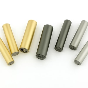 Könnte beinhalten: Fünf verschiedene farbige Metallstempel mit einem geometrischen Muster auf der Oberseite. Die Stempel sind in einer Reihe von links nach rechts angeordnet, wobei der goldene Stempel links und der silberne Stempel rechts ist.