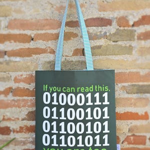 Pode incluir: Bolsa tote verde com texto branco: "Se consegues ler isto, 01000111 01100101 01100101 01101011 tu também". A bolsa tem alças listradas e está pendurada numa parede de tijolos.