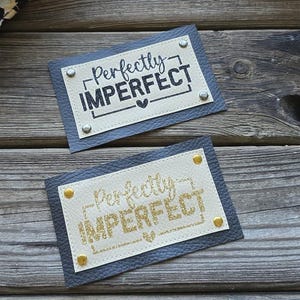Peut inclure: Deux patchs en cuir avec le texte "Perfectly Imperfect" en lettres noires et dorées pailletées. Les patchs sont sur une surface en bois avec un sac à main à imprimé léopard en arrière-plan.