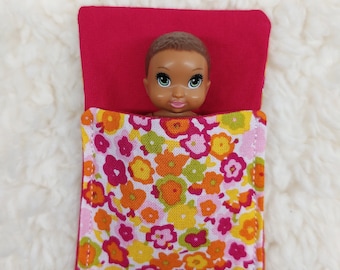 May include: A small doll with brown skin and black hair is lying in a red and white floral sleeping bag.