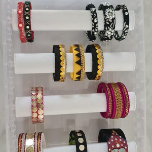 May include: A selection of colourful bangles displayed in a clear plastic organiser. The bangles feature diverse designs, including floral patterns, gold accents, and vibrant colours like red, black, yellow, and pink. The organiser has multiple levels.