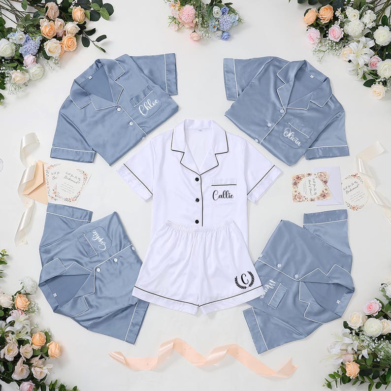 May include: A collection of satin pajama sets in white and blue. The sets include short-sleeved button-down shirts and shorts or pants. Each set is personalized with a name or initial. Floral arrangements and cards are also present.