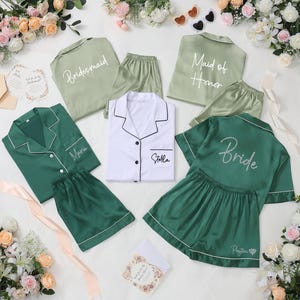 May include: An assortment of satin pajama sets in shades of green and white, with white piping. Some sets are labeled "Bridesmaid", "Maid of Honor", and "Bride". Other sets are personalized with names. The sets are arranged with floral accents.