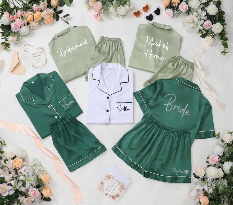 May include: An assortment of satin pajama sets in shades of green and white, with white piping. Some sets are labeled "Bridesmaid", "Maid of Honor", and "Bride". Other sets are personalized with names. The sets are arranged with floral accents.