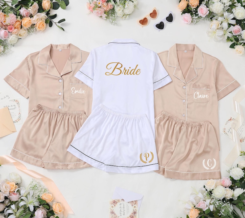 May include: Three sets of satin pajama sets. Two sets are a light tan color, and one is white. The white set has the word "Bride" in gold script on the back. The tan sets have names embroidered on the front pocket.