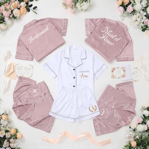 May include: A collection of bridal party pajamas in white and mauve. The pajamas include short-sleeved shirts and shorts or pants. The shirts are embroidered with "Bridesmaid" and "Maid of Honor". The white pajamas are personalized with a name.