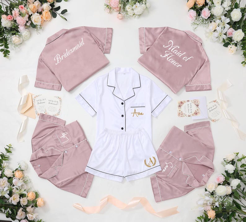 May include: A collection of bridal party pajamas in white and mauve. The pajamas include short-sleeved shirts and shorts or pants. The shirts are embroidered with "Bridesmaid" and "Maid of Honor". The white pajamas are personalized with a name.