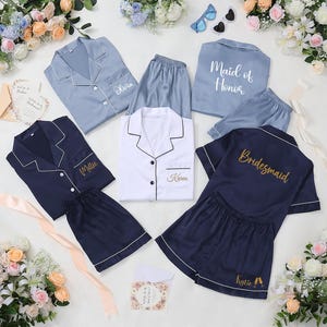 May include: A collection of satin pajama sets in various colors, including light blue, navy, and white. Each set features a button-down top and shorts, with personalized embroidery such as names and titles like "Maid of Honor" and "Bridesmaid".