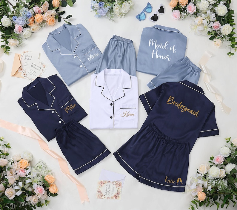 May include: A collection of satin pajama sets in various colors, including light blue, navy, and white. Each set features a button-down top and shorts, with personalized embroidery such as names and titles like "Maid of Honor" and "Bridesmaid".