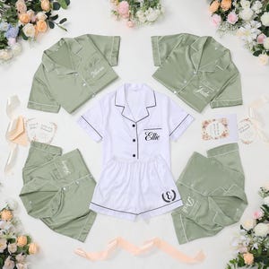 May include: A collection of satin pajama sets in white and sage green, with personalized embroidery. The sets include short-sleeved button-down shirts and shorts or pants. Floral arrangements and invitation cards are arranged around the pajamas.