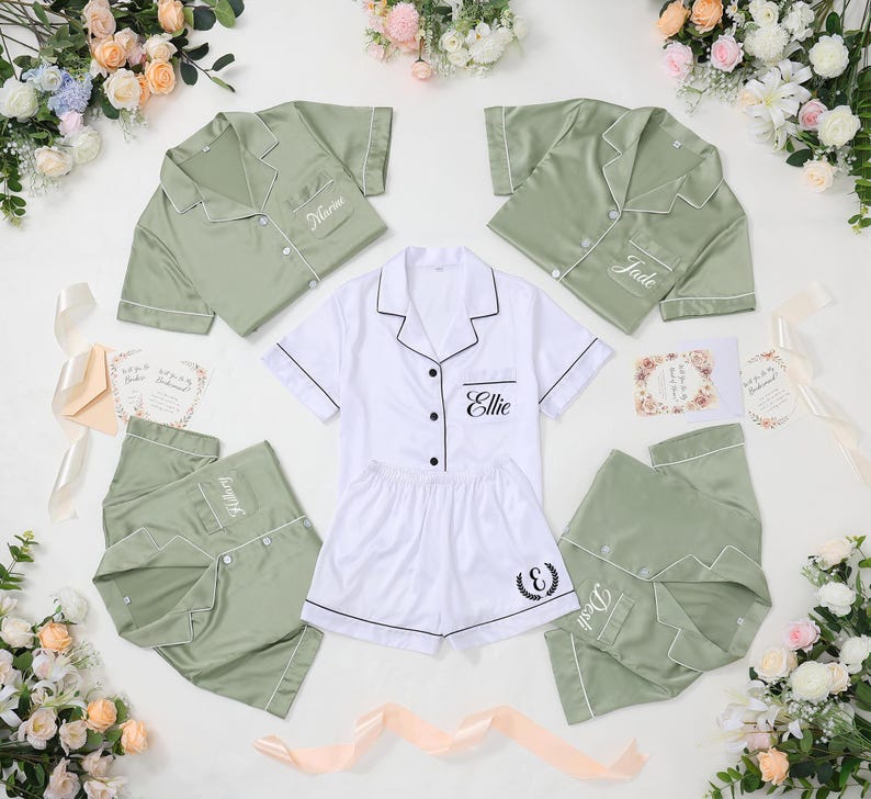 May include: A collection of satin pajama sets in white and sage green, with personalized embroidery. The sets include short-sleeved button-down shirts and shorts or pants. Floral arrangements and invitation cards are arranged around the pajamas.