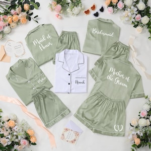 Könnte beinhalten: Satin-Pyjama-Sets in Salbeigrün mit weißem Paspel, personalisiert mit Namen und Titeln wie "Brautjungfer" und "Mutter des Bräutigams". Ein weißes Pyjama-Set mit schwarzem Paspel ist ebenfalls enthalten.