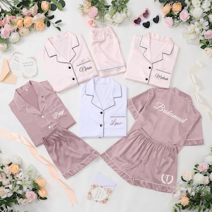 May include: A collection of satin pajama sets in various colors, including pink and white, with black trim. Some sets are personalized with names like "Paige" and "Laura", and the word "Bridesmaid". The sets are arranged with flowers and accessories.