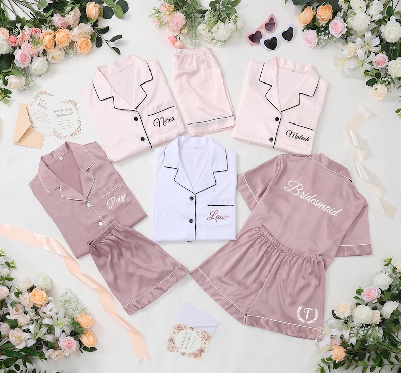 May include: A collection of satin pajama sets in various colors, including pink and white, with black trim. Some sets are personalized with names like "Paige" and "Laura", and the word "Bridesmaid". The sets are arranged with flowers and accessories.