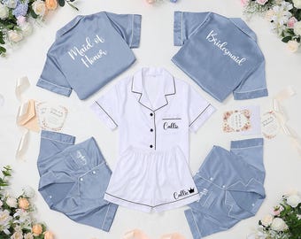May include: A collection of bridal party pajamas in blue and white. The set includes short-sleeved button-down shirts and shorts, with some pieces labeled "Maid of Honor," "Bridesmaid," and names. Floral arrangements and invitation cards surround the pajamas.