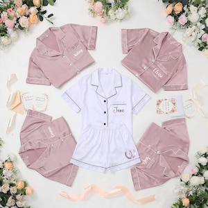 May include: A collection of blush pink and white satin pajama sets, each with personalized embroidery. The sets include short-sleeved button-down shirts and shorts. Floral arrangements and invitation cards are arranged around the pajamas.