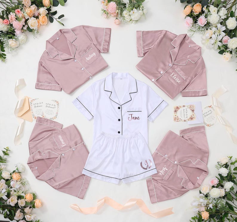 May include: A collection of blush pink and white satin pajama sets, each with personalized embroidery. The sets include short-sleeved button-down shirts and shorts. Floral arrangements and invitation cards are arranged around the pajamas.