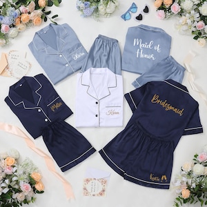 May include: A collection of satin pajama sets in various colors, including light blue, navy, and white. Each set features a button-down top and shorts, with personalized embroidery such as names and titles like "Maid of Honor" and "Bridesmaid."