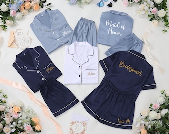 May include: A collection of satin pajama sets in various colors, including light blue, navy, and white. Each set features a button-down top and shorts, with personalized embroidery such as names and titles like "Maid of Honor" and "Bridesmaid."