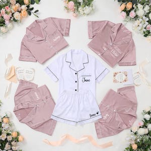 May include: A collection of satin pajama sets in white and dusty rose. Each set includes a button-down shirt and shorts, with personalized names embroidered on the pocket and shorts. The pajamas are arranged with floral accents and invitation cards.