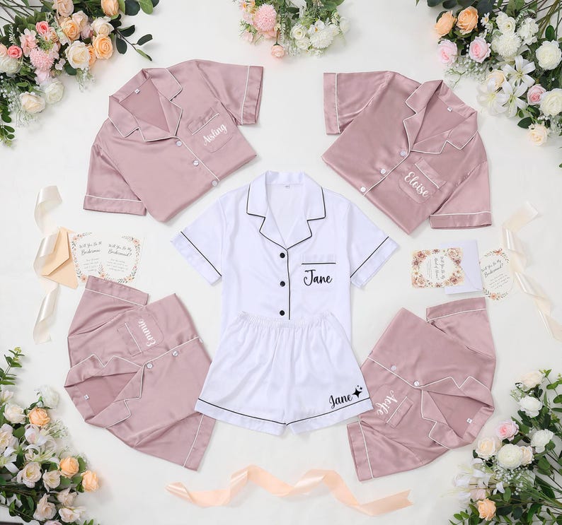 May include: A collection of satin pajama sets in white and dusty rose. Each set includes a button-down shirt and shorts, with personalized names embroidered on the pocket and shorts. The pajamas are arranged with floral accents and invitation cards.