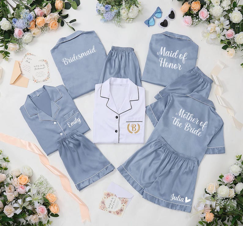 May include: A collection of light blue satin pajama sets, including tops and shorts, with white piping. Some sets are labeled "Bridesmaid," "Maid of Honor," "Mother of the Bride," and "Emily" with a gold embroidered initial. The image is decorated with flowers and stationery.