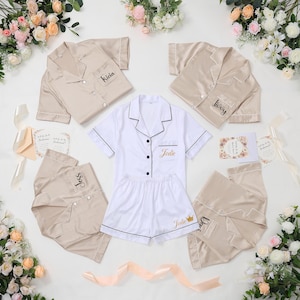 May include: A collection of personalized satin pajama sets in cream and white. Each set includes a button-down shirt and shorts or pants, with names embroidered on the shirt pocket. Floral arrangements and cards surround the sets.