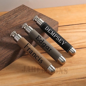 Könnte beinhalten: Drei personalisierte Flachmänner mit Edelstahlakzenten und Lederhüllen. Die Flachmänner sind in den Farben Beige, Grau und Schwarz gehalten, jeder mit einem weißen Namensaufdruck. Die Flachmänner werden auf einer Holzoberfläche präsentiert.