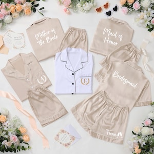 May include: A collection of beige and white satin pajama sets, some with text like "Mother of the Bride," "Maid of Honor," and "Bridesmaid." Each set includes a button-down shirt and shorts. Floral arrangements and invitation cards surround the sets.