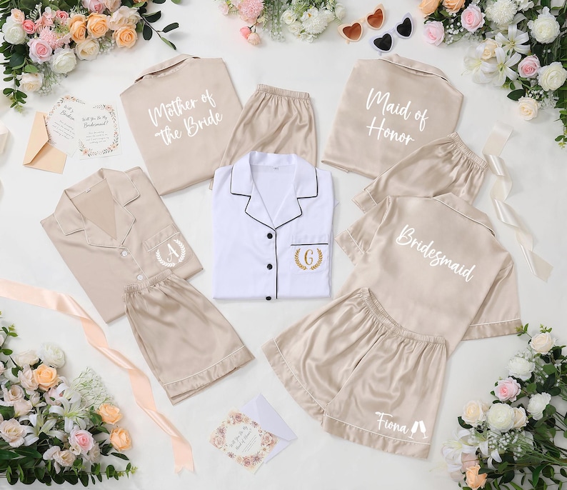 May include: A collection of beige and white satin pajama sets, some with text like "Mother of the Bride," "Maid of Honor," and "Bridesmaid." Each set includes a button-down shirt and shorts. Floral arrangements and invitation cards surround the sets.