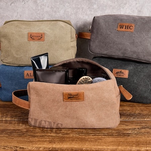 May include: A collection of canvas toiletry bags in various colors, including tan, gray, and blue. Each bag features a leather handle and a personalized leather patch. One bag is open, revealing grooming products.