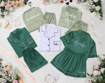 May include: An assortment of satin pajama sets in shades of green and white, with white piping. Some sets are labeled "Bridesmaid", "Maid of Honor", and "Bride". Other sets are personalized with names. The sets are arranged with floral accents.